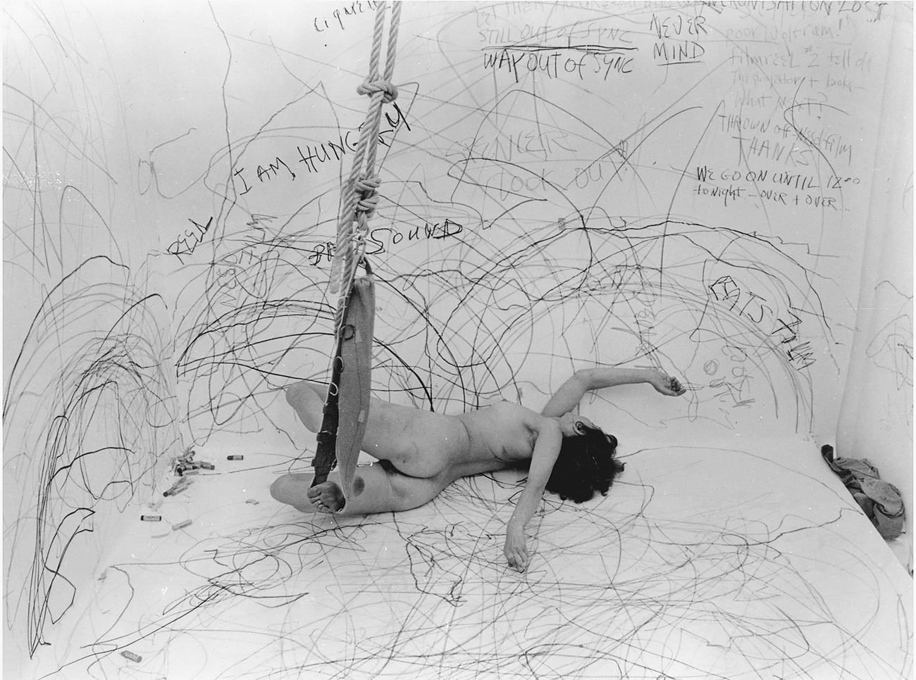 Carolee Schneemann lying on the floor of a room with chaotic scribbles and text on the walls.