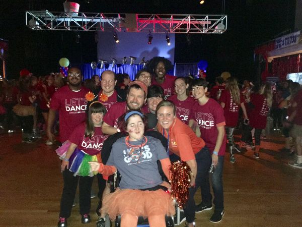 A group of eleven people gather in a dance hall. Many wear Let�s Dance Ottothon 2017 T-shirts. A woman in the center wears a Life Is Good T-shirt.