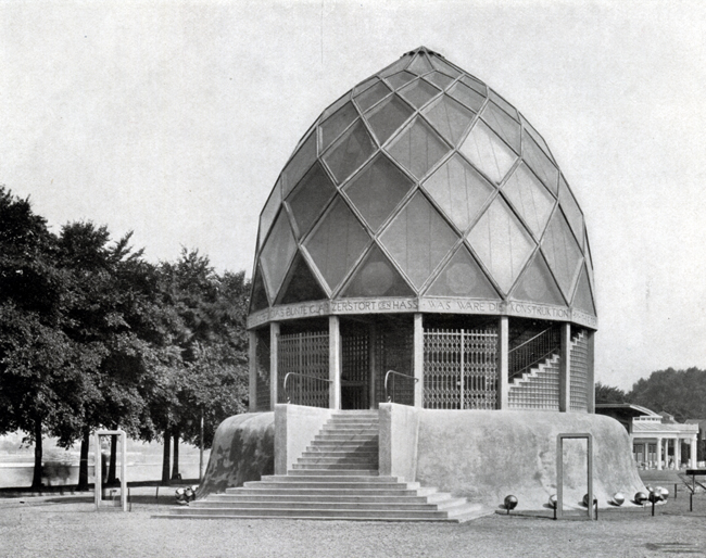 The exhibit showcases Bruno Taut’s Glashaus exterior at the Cologne Werkbund exhibition from a distance. The structure has a dome-shaped roof with a steel-reinforced body, and the entrance features stairs. Trees can be seen in the background.