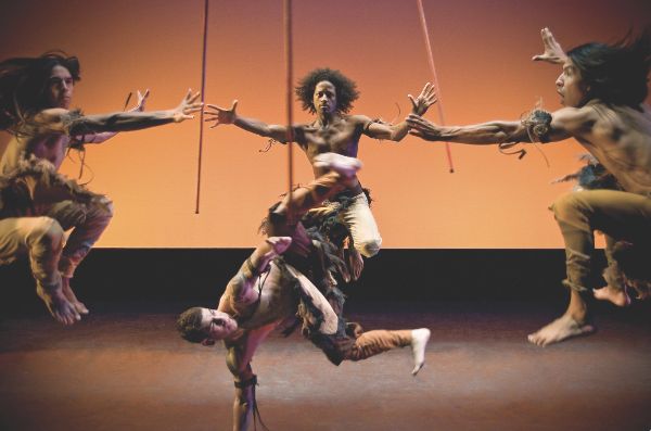 Three bare-chested dancers jump high, knees bent, arms out, three sticks hovering between them. In center dancer balances on his hand, kicking out.