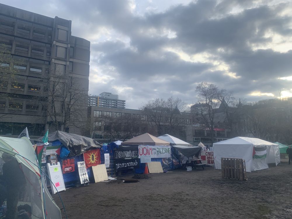On a cloudy day, tents are pitched on a dirt clearing in the UAQM neighborhood in Montréal. Posters are pinned to the tents and concrete university buildings are seen in the background.