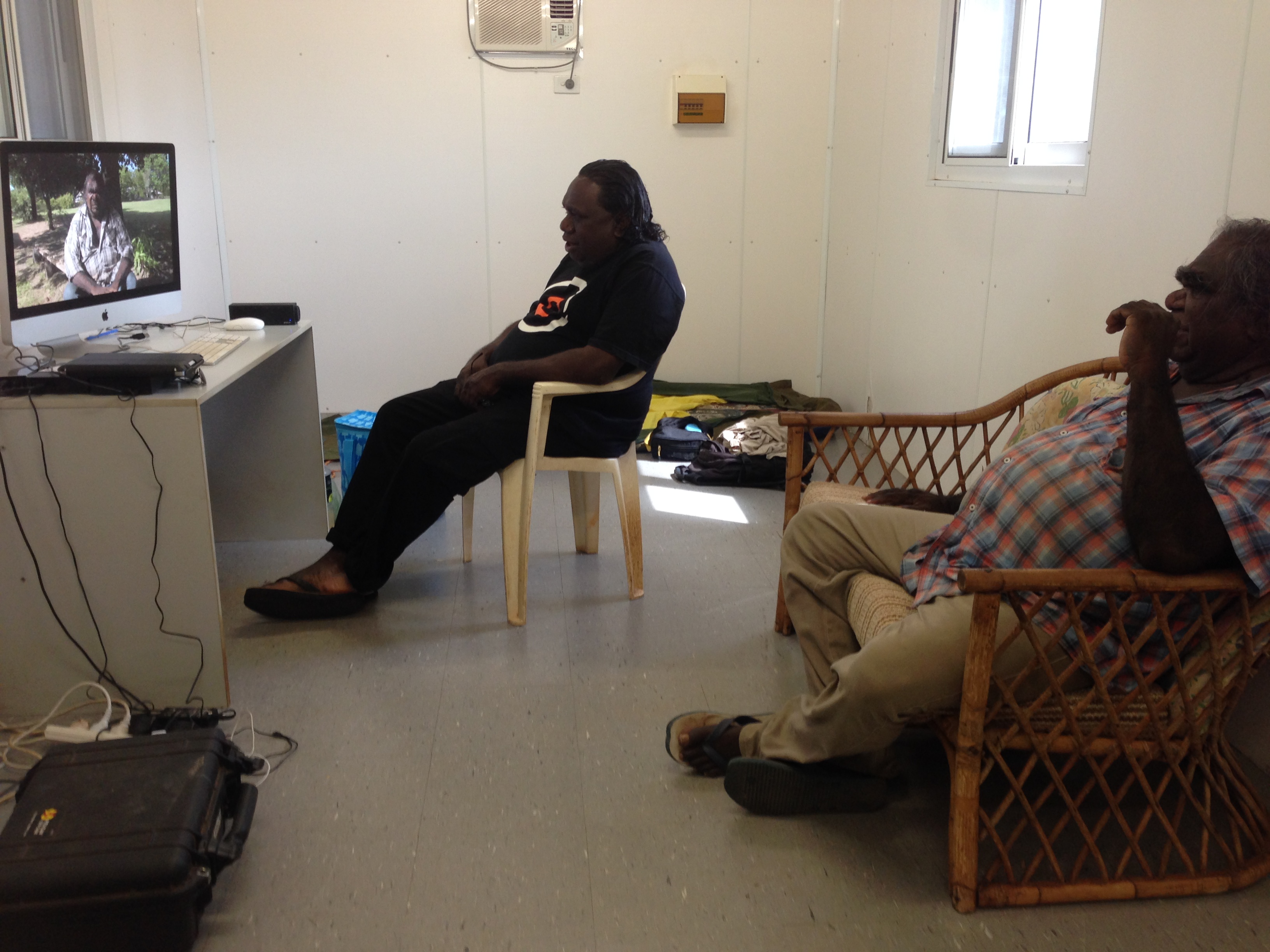 In a white-walled room, Dunba and Henry are sitting in chairs viewing a computer screen displaying Dunba’s film on the left side.