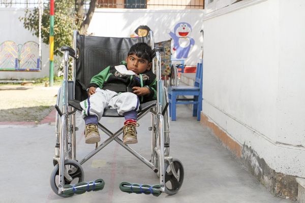 A young Indian disabled child sits in a large manual wheelchair, leaning to the left against a nonergonomic seat back with his legs dangling.