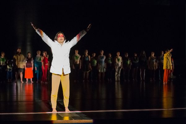 A dancer wearing a white parka stands with arms stretched up and out. Behind her, on the dark stage, a group of people is walking around.