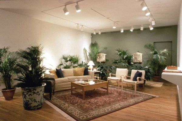 An empty living room with a couch, two chairs, a number of potted plants, two bird houses, coffee table, and center rug.