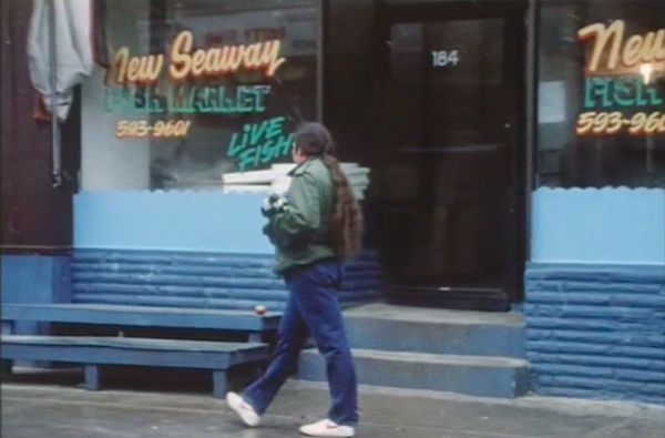 Thomas walks in front of blue benches and a storefront. The painted letters on the window state “New Seaway Fish Market—Live Fish—593-9601.”
