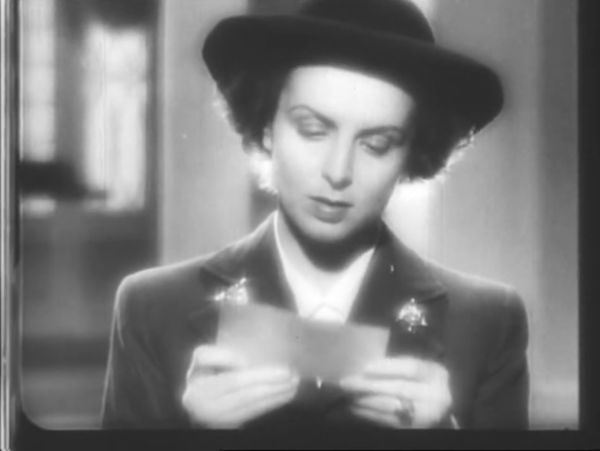 A woman dressed in fine clothing with a hat and jacket intently looks down to read a letter held in her hand.