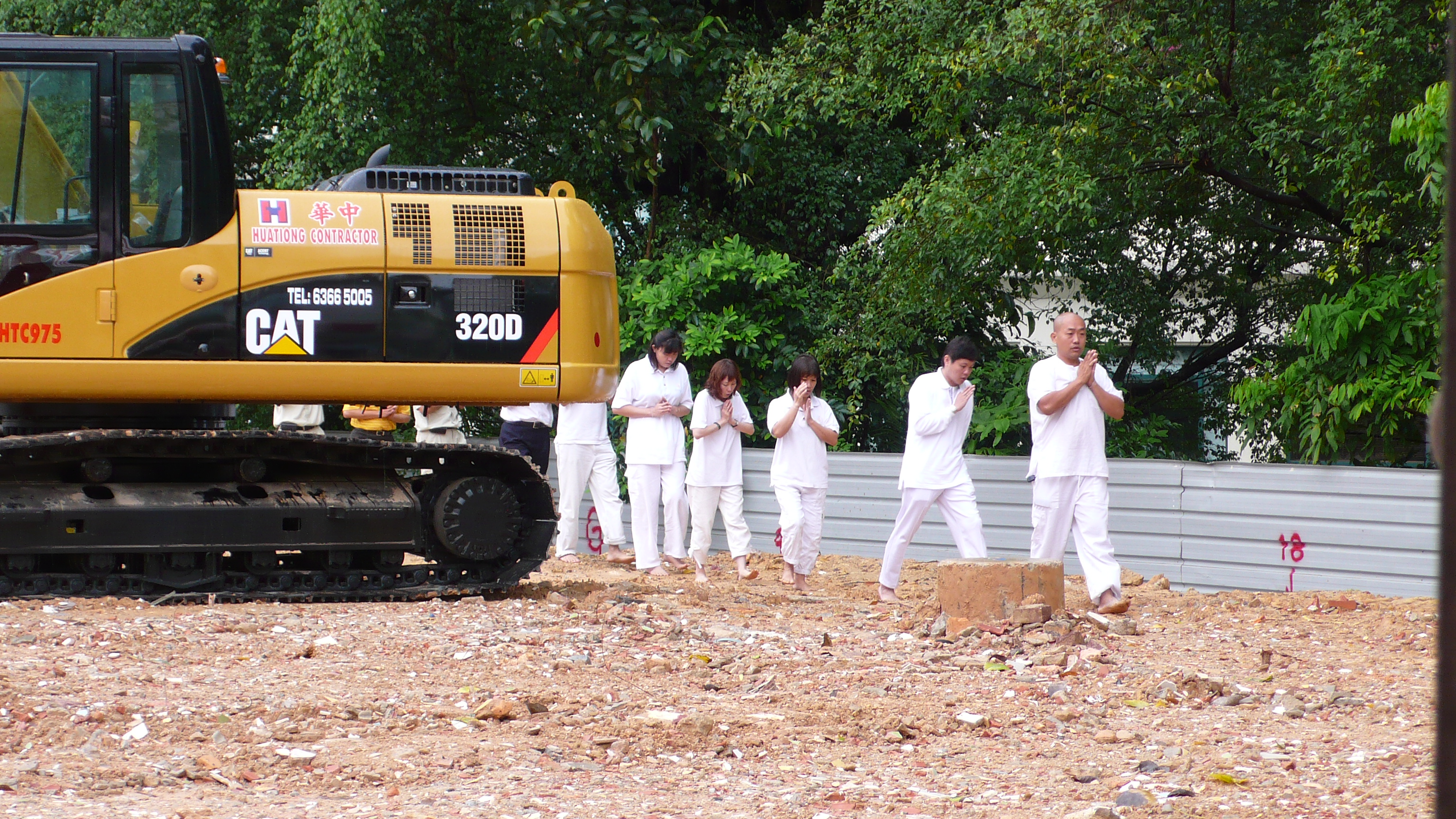 A row of barefoot men and women in white walk at the edge of a muddy site with their hands in prayer position. An excavator is at left.