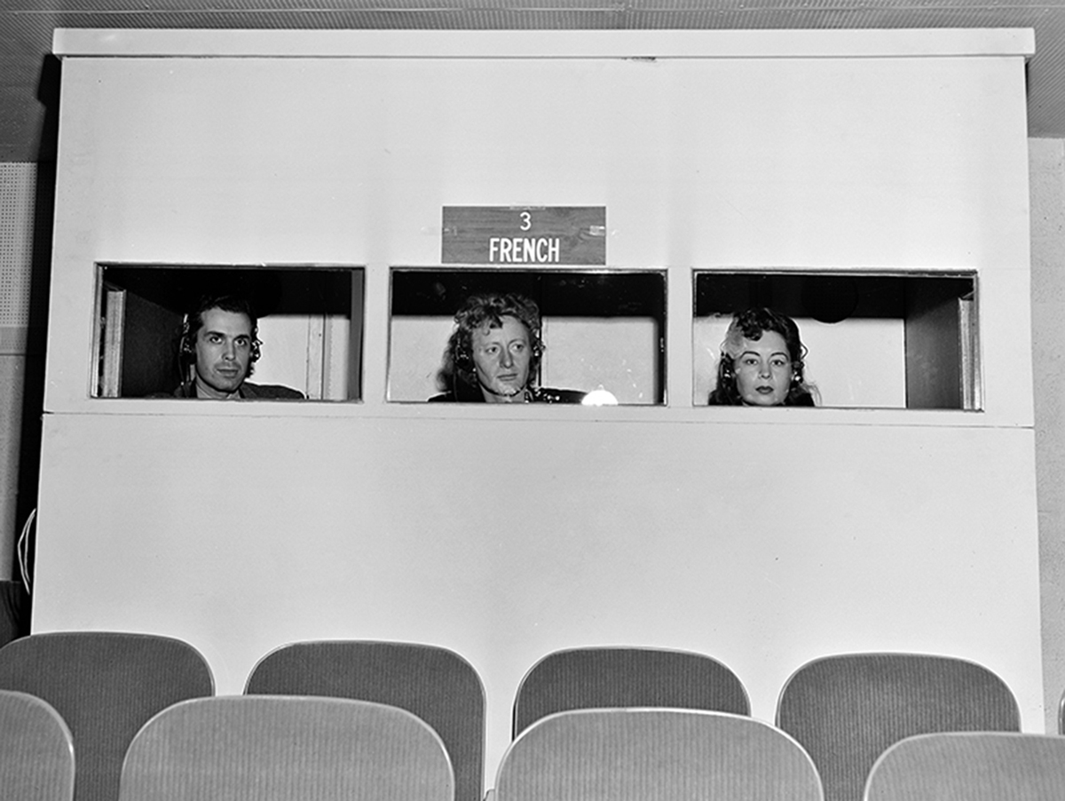 Photograph of interpreters’ booth showing one male and two female interpreters wearing headphones under a label reading “3 French.”
