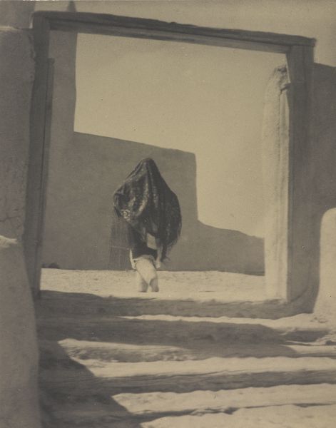 A woman in a loosely draped shawl walks through a square wooden portal at the top of shallow stairs, toward an adobe building.