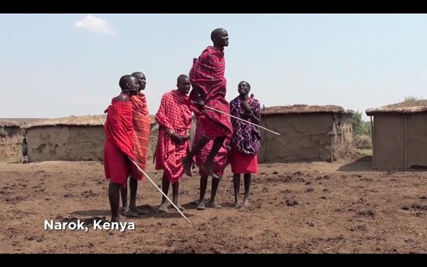 Six men in front of thatched structures, one caught mid-jump