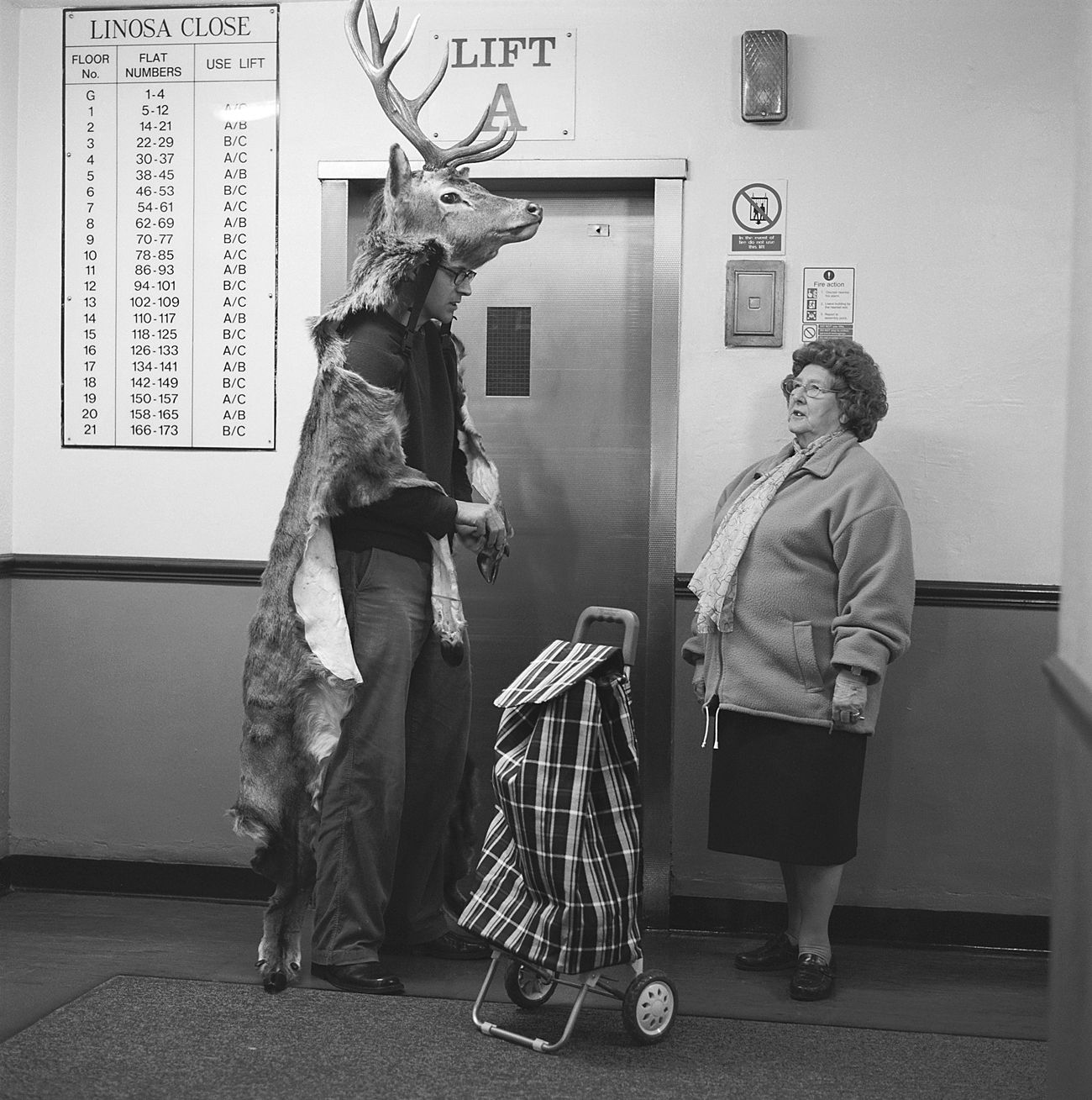 Marcus Coates wearing a deer costume stands near an elevator, speaking to an older woman with a shopping trolley in a residential building.