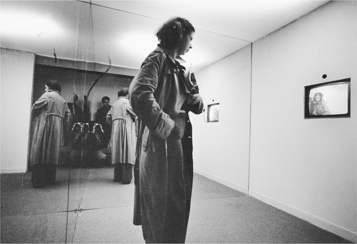 A photograph of a woman in a room with 2 sides of it mounted with mirror walls. The woman is staring at a time-delayed image of herself on the monitor placed on one side of the wall.