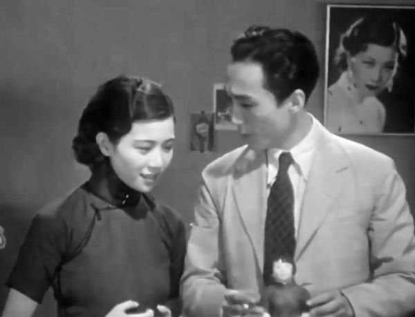A couple stand side by side with a doll before them and the funeral portrait from the previous image on the wall behind them.