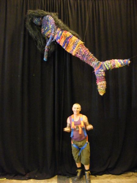 Two figures in front of a black theater curtain: a person in a climbing harness looking at the camera; suspended from above a fabricated creatue with a human torso and a fish tail instead of legs.