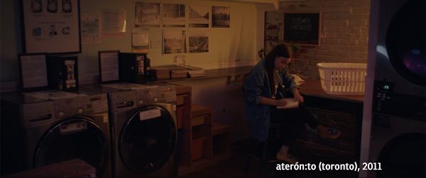 Yellow-hued image of laundromat. A Mohawk woman with long brown hair is writing in a journal. The bottom right states “aterón:to (toronto), 2011.”