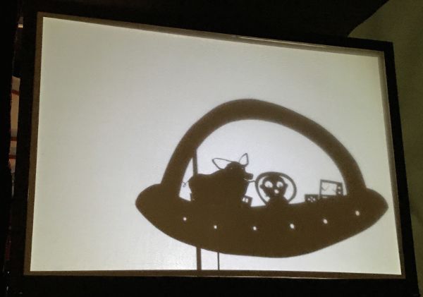 Shadow puppet of a flying saucer with two occupants: a human and a pig.