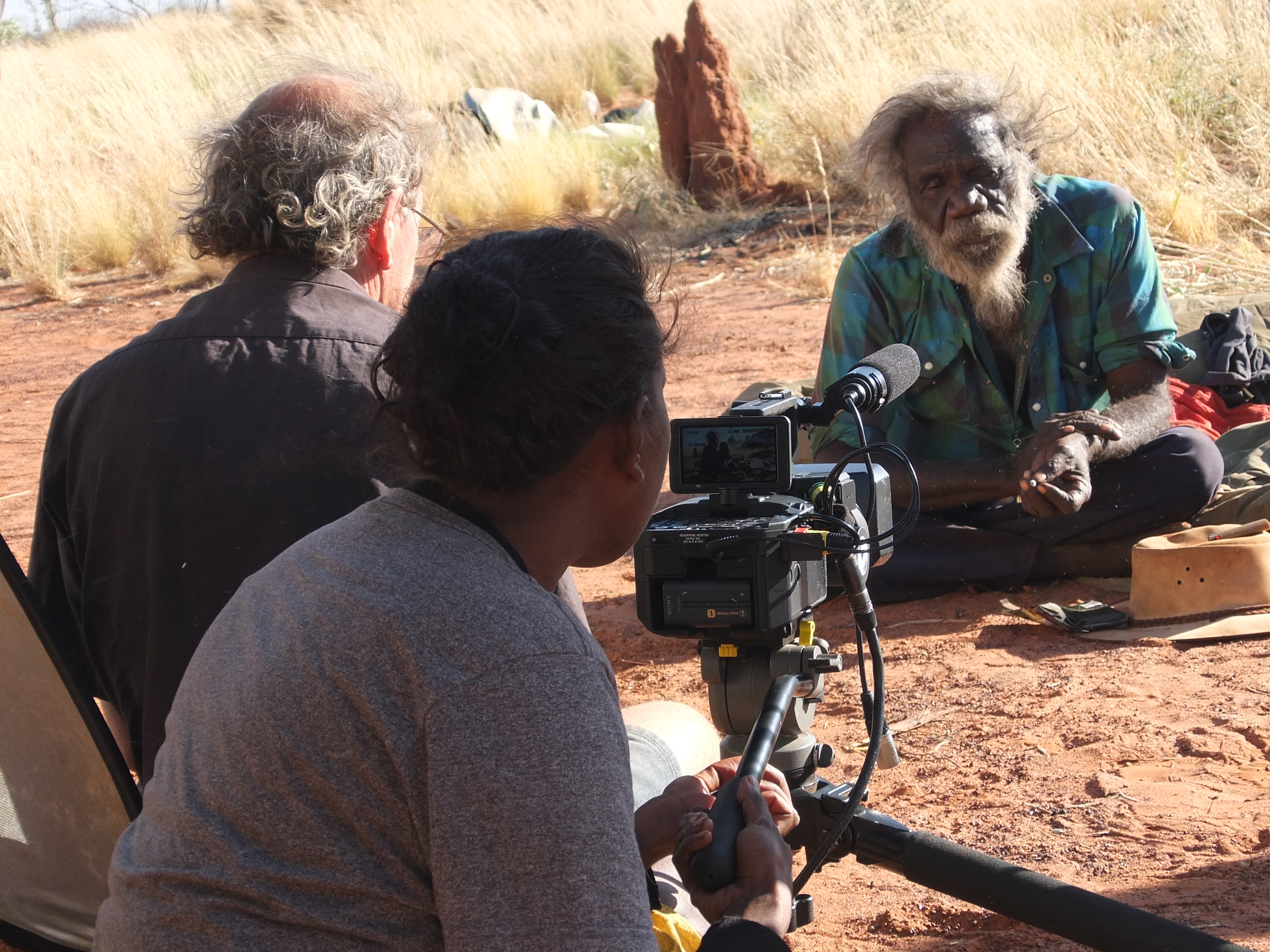 Mark is sitting in front of a termite mound, speaking into the camera in the foreground. Anne is adjusting it and Neil sits to the left.