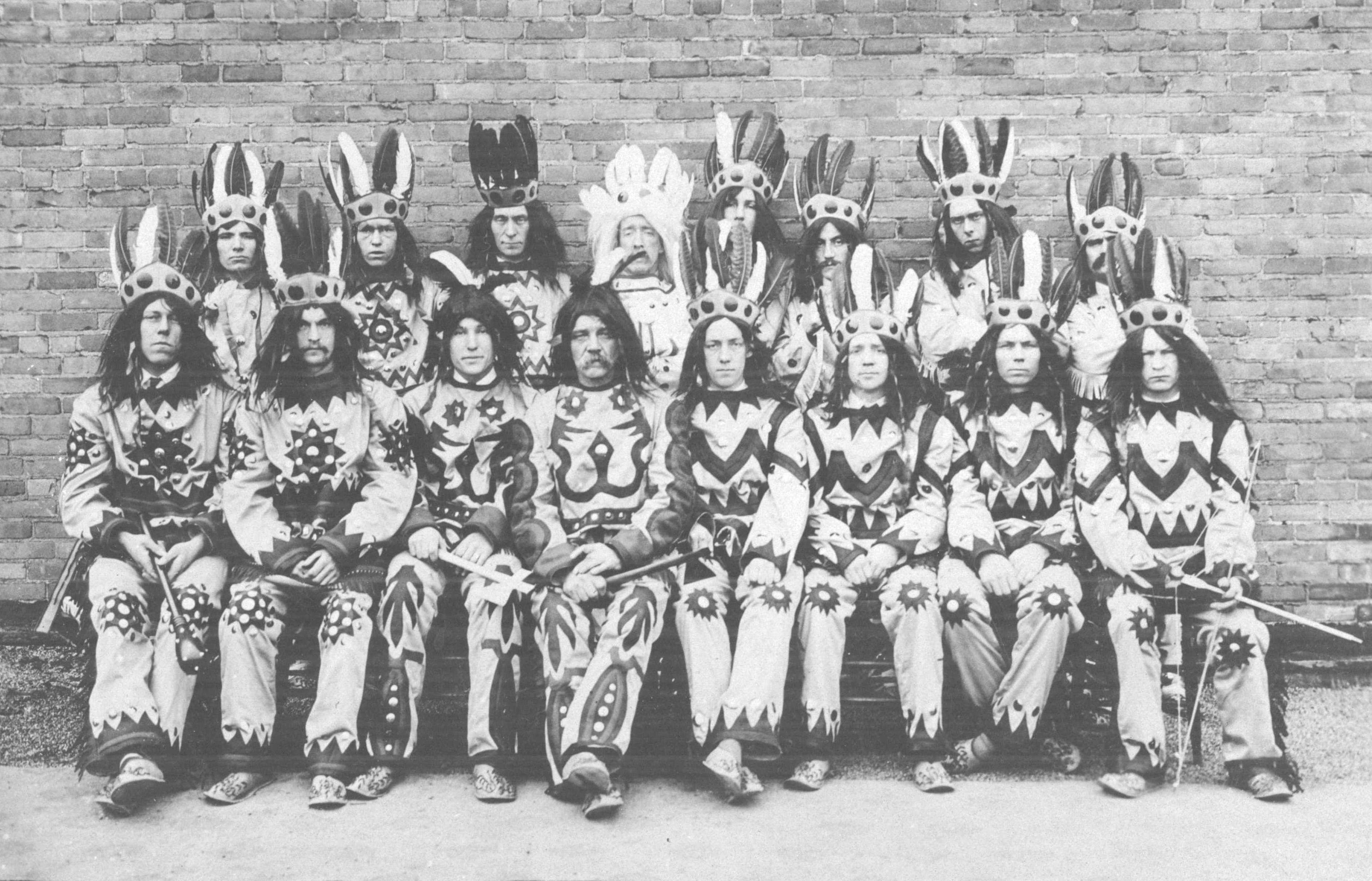 Two rows of white men are posed wearing Native American costumes with headdresses. Some are holding Native American weapons or hunting tools.