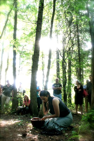 In a forest with tall trees, light pouring in, a woman kneels before a bowl, one hand in it and the other lifted to her hair. A group of people watch.