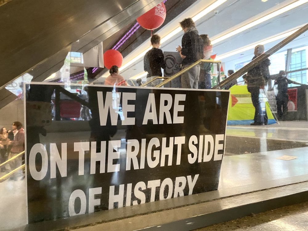 Against a glass divider at The New School is propped a poster that reads “We are on the right side of history.” Student protestors are seen in the background of the photo near pitched tents.