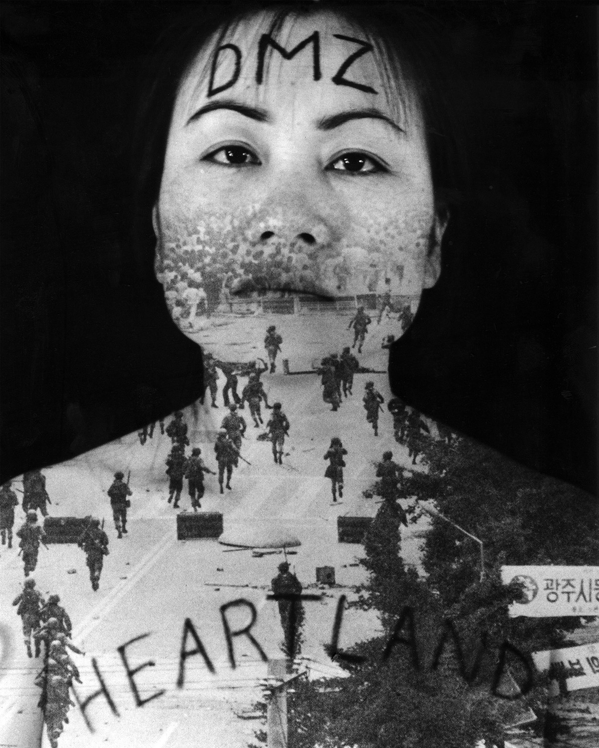 A woman standing in front of a black backdrop with am image of armed soldiers moving down a street projected onto her torso. Written in ink across her forehead are the letters DMZ and across her chest the word Heartland
