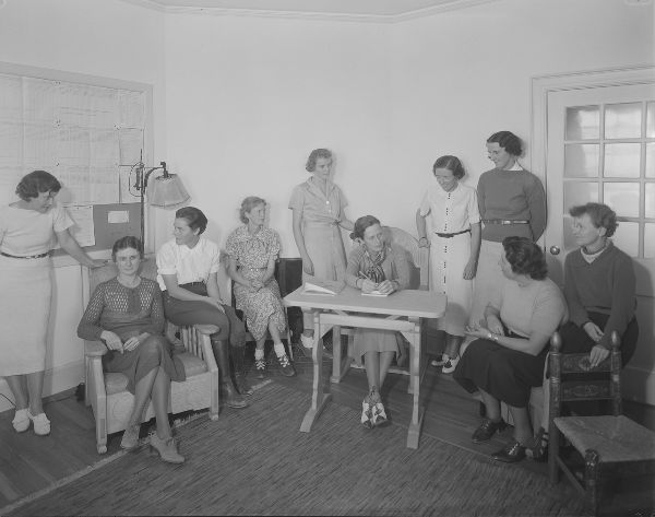 Ten women are seated casually in a classroom, conversing. All wear dresses or skirts except for one in jodhpurs.