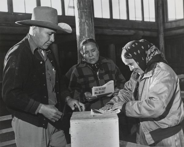 A man in a broad-brimmed hat looks on as two women prepare to place their ballots in a ballot box.