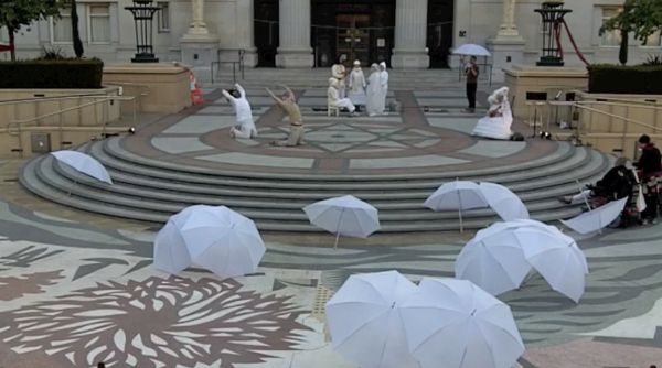 A plaza with white umbrellas laying in the foreground and performers in early twentieth-century dress in the background.