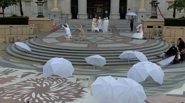 A plaza with white umbrellas laying in the foreground and performers in early twentieth-century dress in the background.