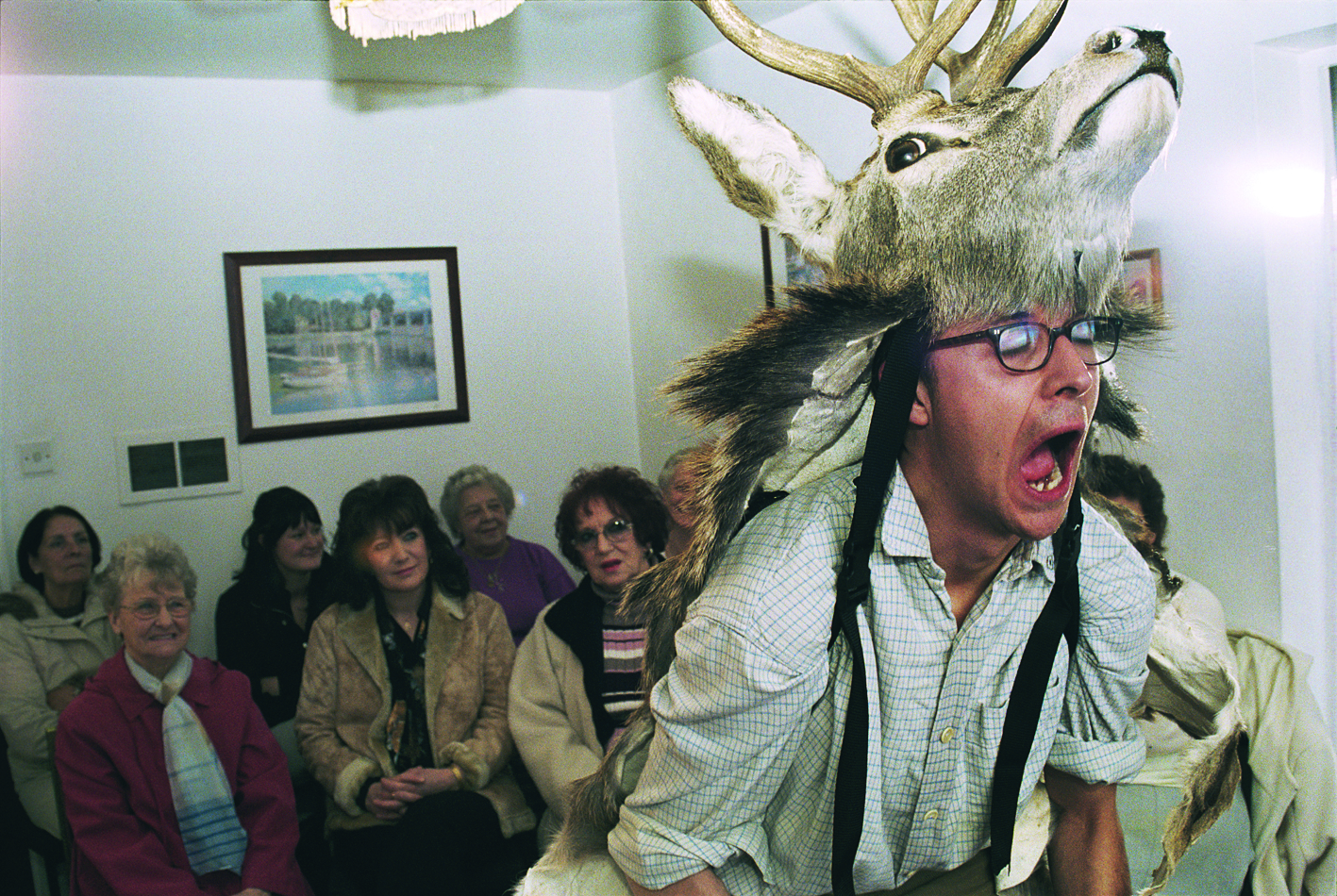Marcus Coates wearing a deer head costume performs theatrically in front of an audience seated in a small room.