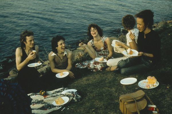 Five women make merry while eating cake off of paper plates and sitting on the grass; behind them is a line of rocks that borders a river.