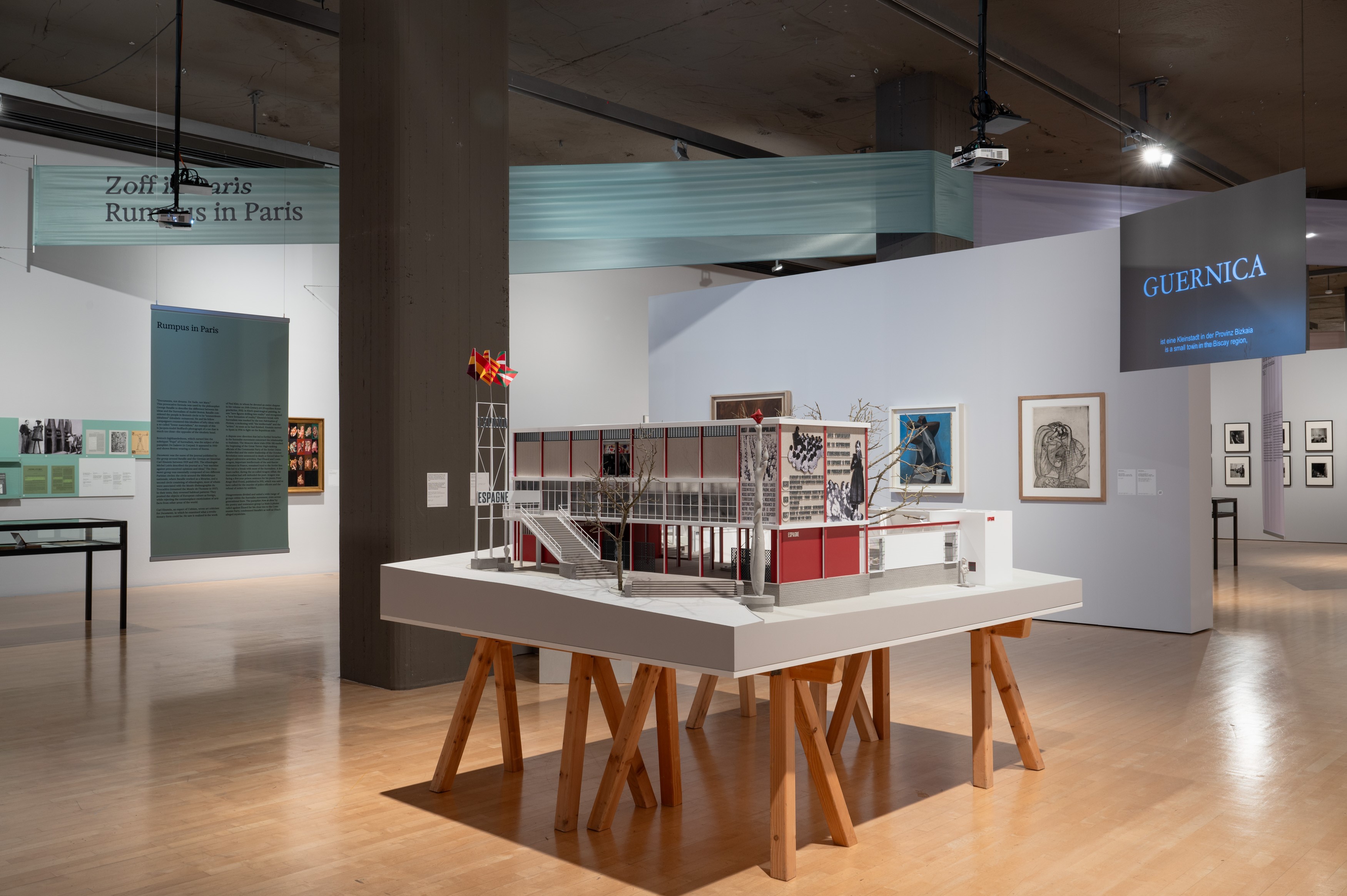 Interior shot of the exhibition space, featuring a model of the Spanish Pavilion in the centre which sits stop a white table with wooden legs. Art works hang on the walls and gallery text and labels are suspended from the ceiling or wrapped around pillars.