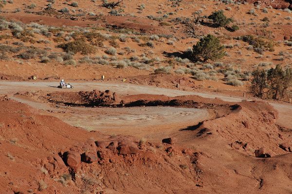 A road through Monument Valley, seen from above. A small figure in a vehicle travels from left to right.