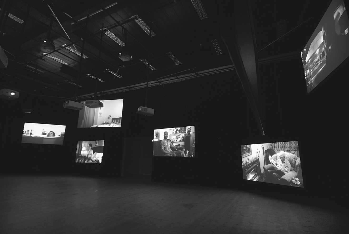 A dark exhibition space featuring multiple video projections on dark walls, each showing scenes of people in domestic settings.