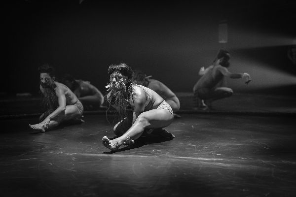 Four dancers bend low, left legs out straight, gazes intense, beard-like straw-looking things hanging from their faces. Man behind crouches, arms out.