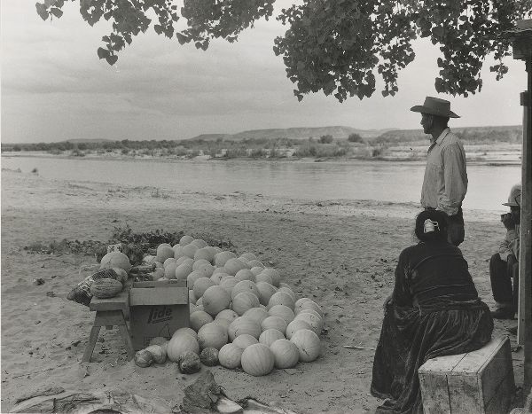 A heap of melons next to a bench on a riverbank. Three people sit or stand in the shade of a tree on the right, all looking away from the camera.