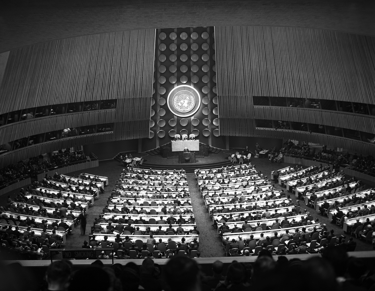 A photo of four curved ranks of seats, filled with people. A curved wall of wooden mullions framing the UN symbol embraces the speaker facing them.