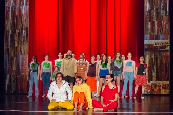 Two long strips with fish photos and the word Que�ana hang. Between, three dancers sit in front, looking out. Behind them, two rows of people stand.