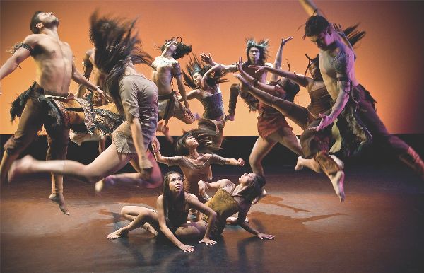 Large group of dancers jump up, hair flying, many arching back. Three women are on the ground in the center, looking out and starting to lift up.