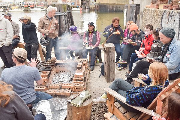 People sit on a loading dock by a river, around a fire pit made of stacked bricks, talking and eating. Smoke is rising and something is cooking.