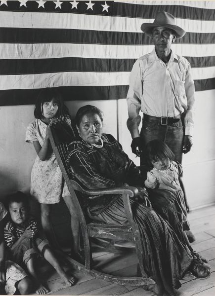 A close-up of a man standing behind a seated woman and four children, in front of a large United States flag hanging on the wall.