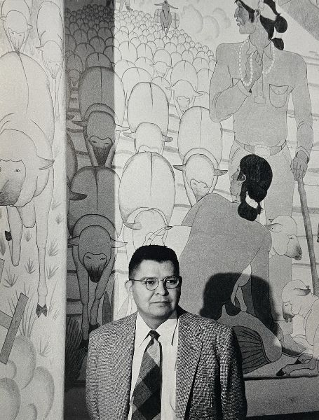A man in a suit and tie, seen from the waist up, stands in front of a mural depicting shepherds with a flock of sheep.