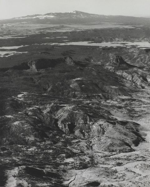An aerial view of foothills in the foreground and a partially snow-covered peak on the horizon.