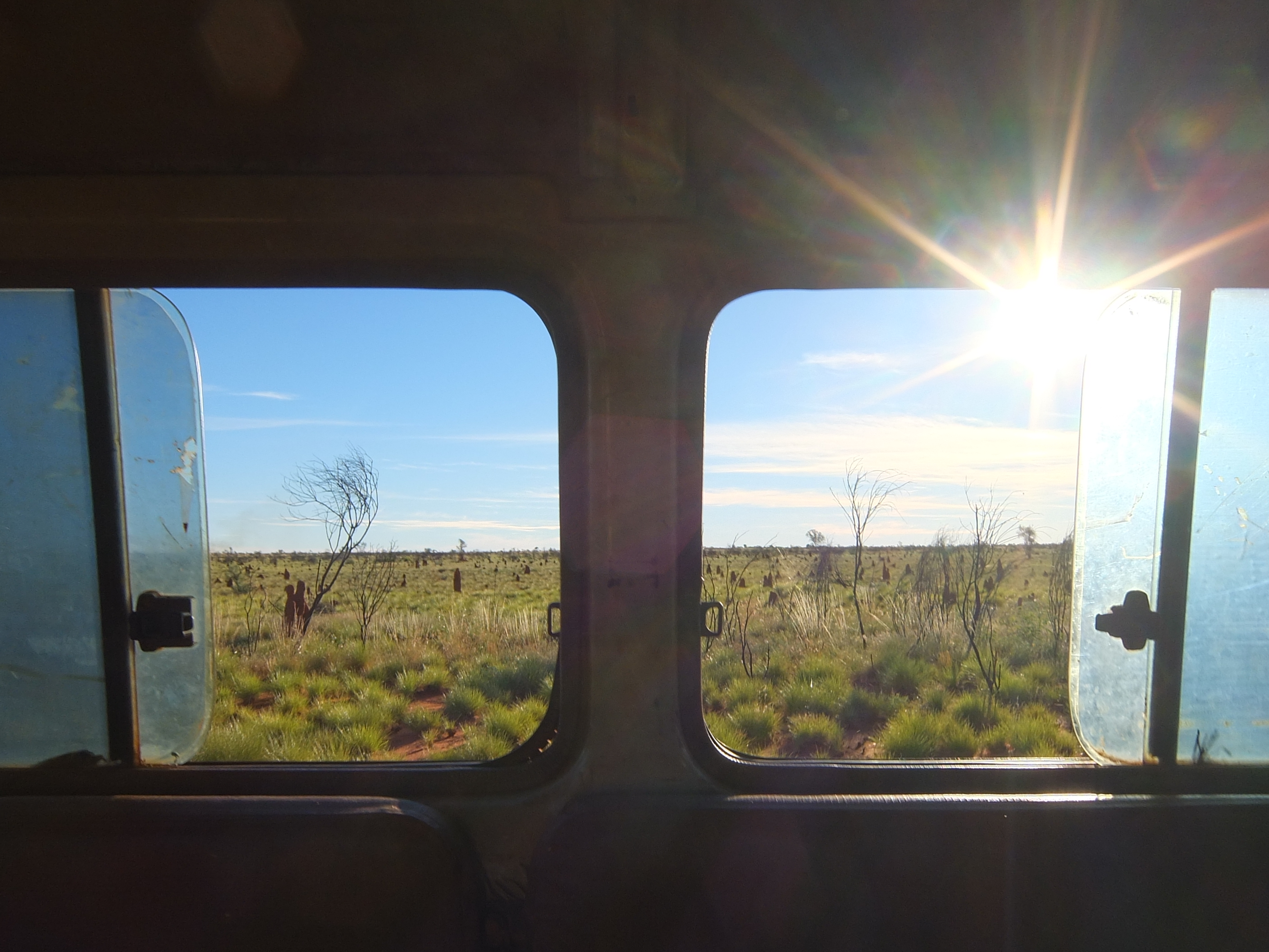 A view through two open windows out of the side of a troopy, showing green desert grasses, a blue sky, and a sun creating a lens flare.