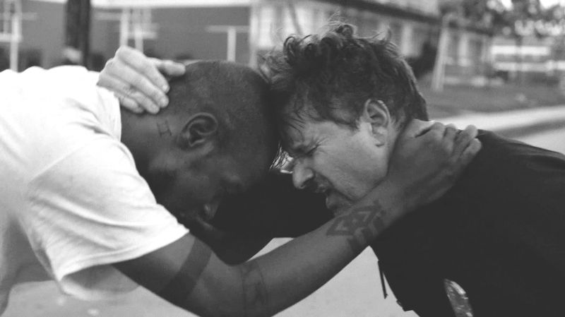 A black man and a white police officer grapple with one another, exhausted, bent over, heads together, and arms around the shoulders of the other.
