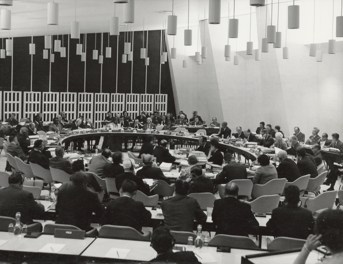 Photograph of delegates convening around an oval table with lights in concentric arrangements suspended from the ceiling above.