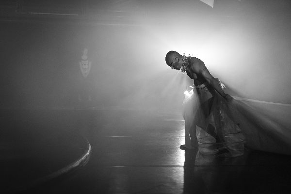 Bald, bare-chested dancer shown sideways, holding long skirt train, walks forward onto a misty stage, leaning in with knees bent, head bowed, pulling.