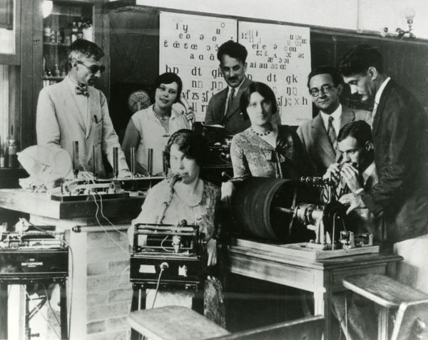 Five men and three women in a room clustered with desks and scientific instruments like early audio recording devices