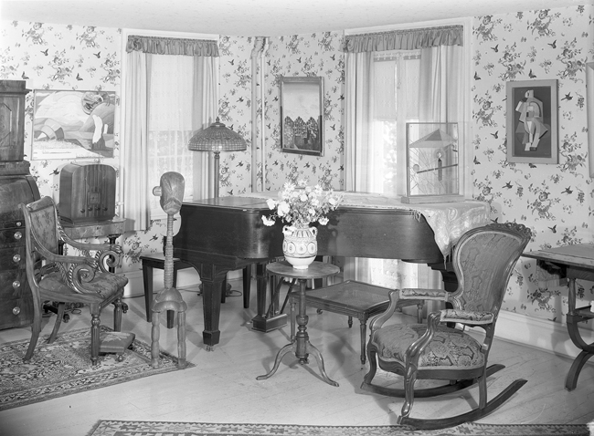 The exhibit displays a corner of a well-furnished room in Katherine S. Dreier’s West Redding home, known as “The Haven.” The scene includes a piano and features artworks such as Constantin Brancusi’s “Little French Girl” (Guggenheim), Max Ernst’s “Paris rêve,” and Marcel Duchamp’s “Small Glass.”
